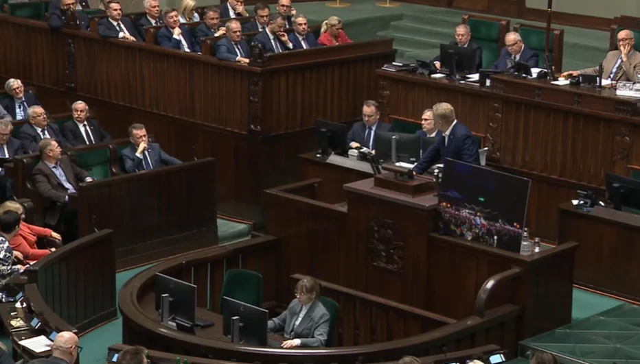 Mówca stojący przy mównicy w sali sejmu, otoczony siedzącymi posłami oraz urzędnikami, skupionymi na wystąpieniu.