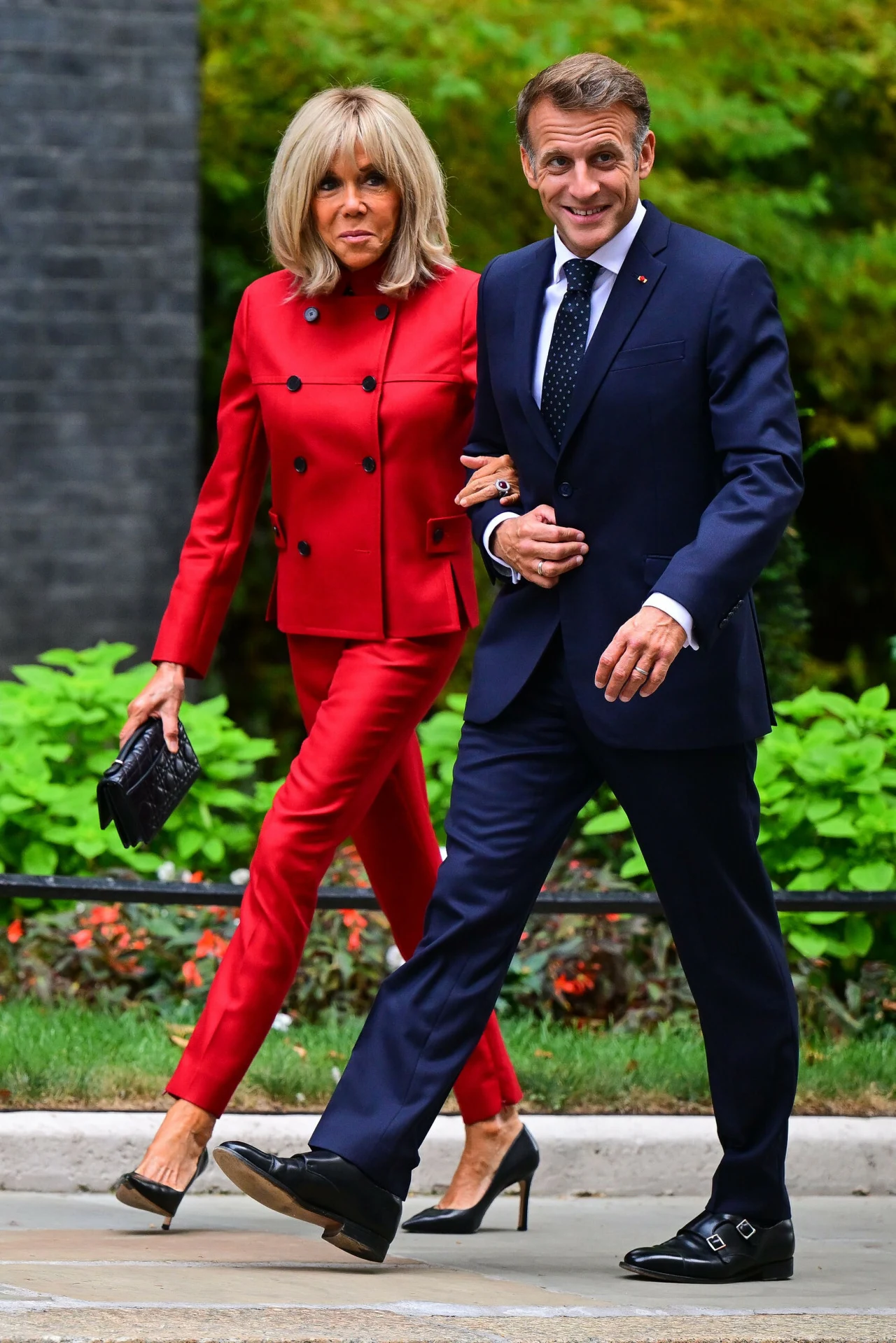 Brigitte Macron zachwyciła w czerwonym garniturze. Wyrazisty kolor skradł show Elegancka kobieta w czerwonym garniturze i czarnych szpilkach idzie pod rękę z mężczyzną w ciemnogranatowym garniturze, koszuli i krawacie; oboje uśmiechnięci podczas spaceru, otoczeni zielenią roślin.