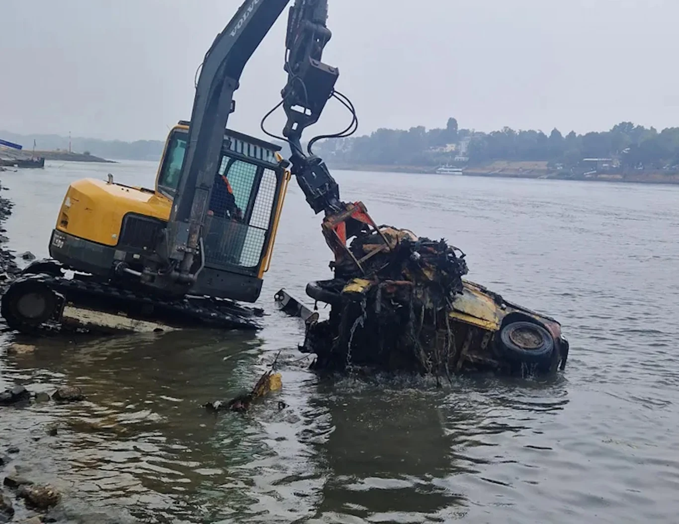 Koparka wyciąga z wody poważnie zniszczony wrak samochodu przy brzegu rzeki.
