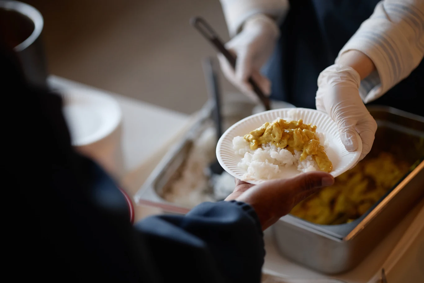 Dieta planetarna ma wspierać zdrowie uczniów w szkołach. Jakie są jej założenia? Osoba w rękawiczkach podaje porcję ryżu z sosem do plastikowego talerza trzymanego przez drugą osobę.