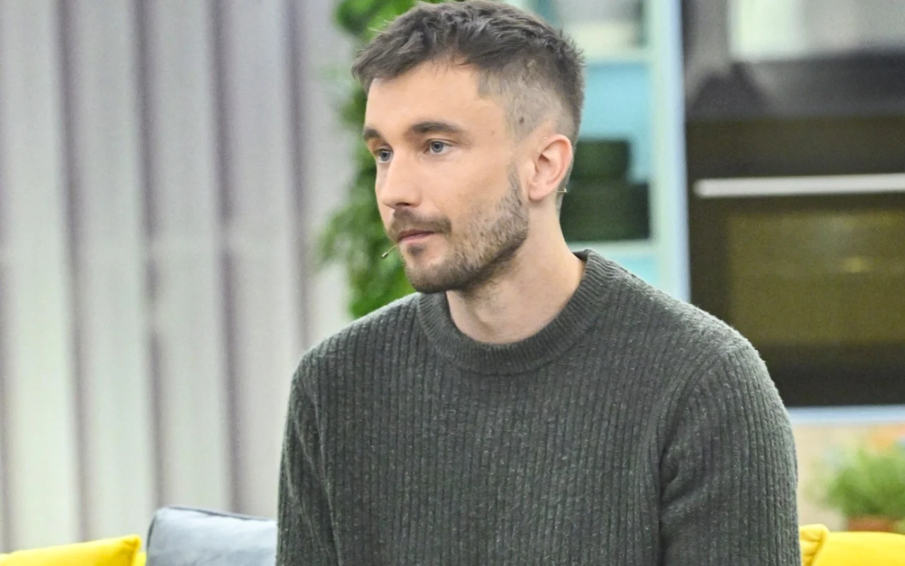 Arkadiusz Wilczyński Mężczyzna z krótkimi, ciemnymi włosami i zarostem, ubrany w ciemnoszary sweter, siedzi na sofie w jasnym pomieszczeniu.