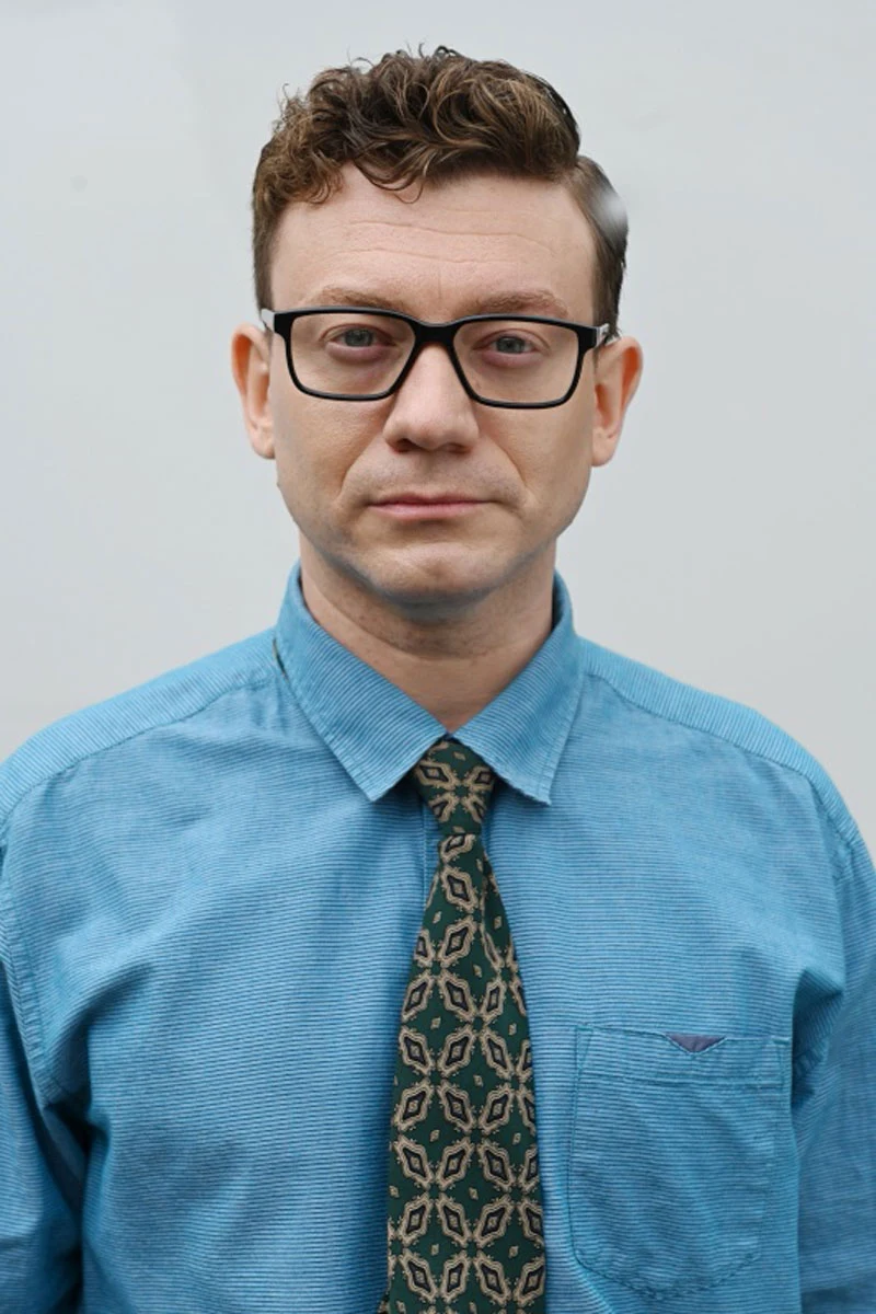 Krzysztof Szczepaniak jako Kamil Wichrowski w "Komisarzu Aleksie" Mężczyzna w okularach i niebieskiej koszuli z wzorzystym krawatem patrzy poważnie wprost przed siebie na jasnym tle.