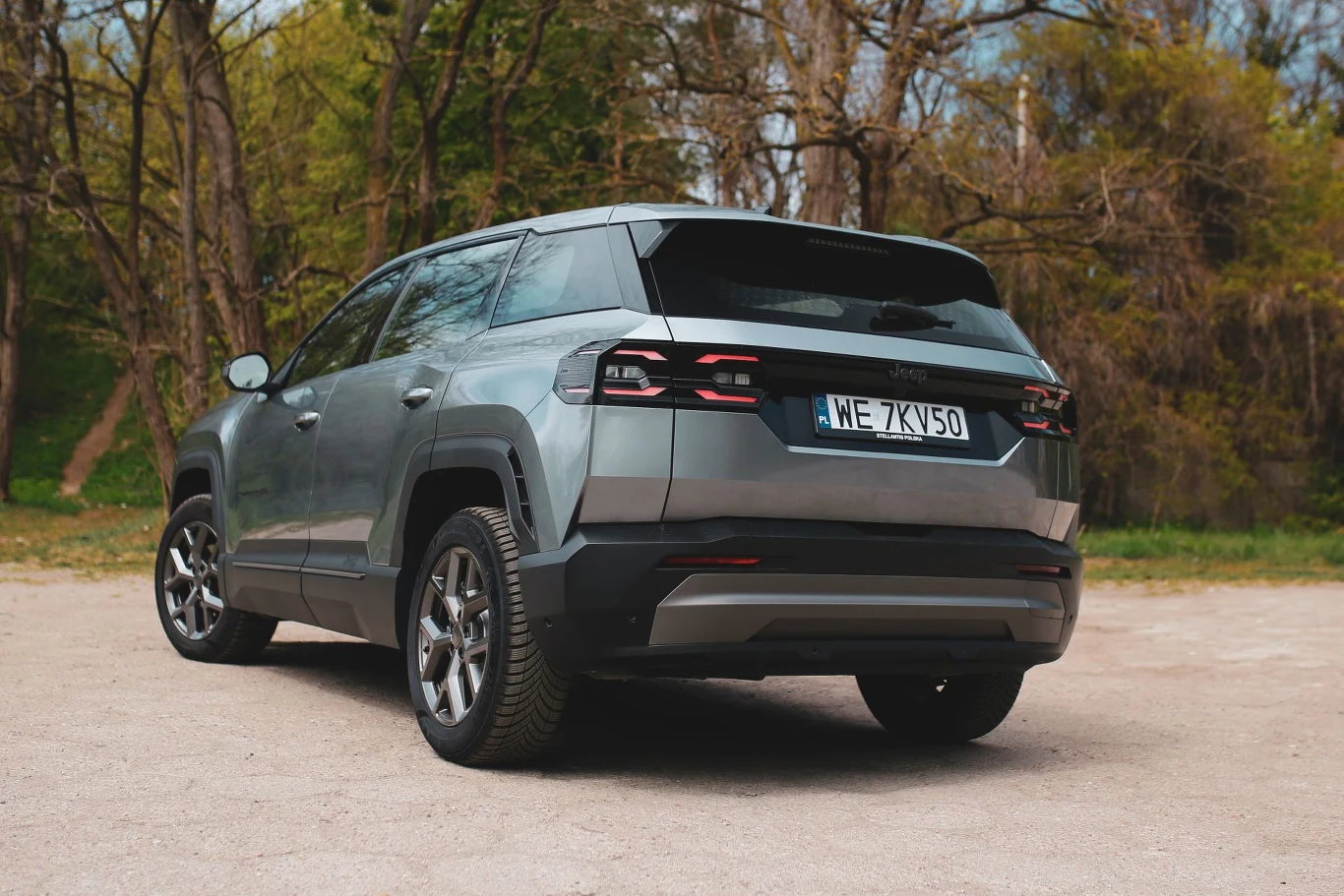 Jeep Compass trzeciej generacji jest o prawie 15 cm dłuższy od swojego poprzednika. Srebrnoszary SUV o nowoczesnym, kanciastym designie, widoczny od tyłu, stoi na żwirowej nawierzchni pośród drzew bez liści.