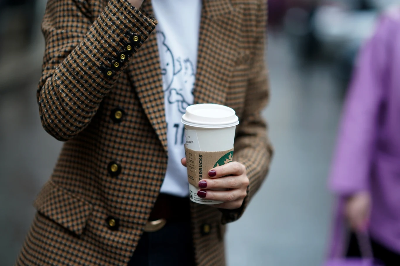 Stylowy manicure w kolorze kawy powraca. Jesienią to idealna propozycja dla minimalistek Osoba w kraciastej marynarce trzyma papierowy kubek kawy Starbucks, eksponując dłonie z bordowym manicure.