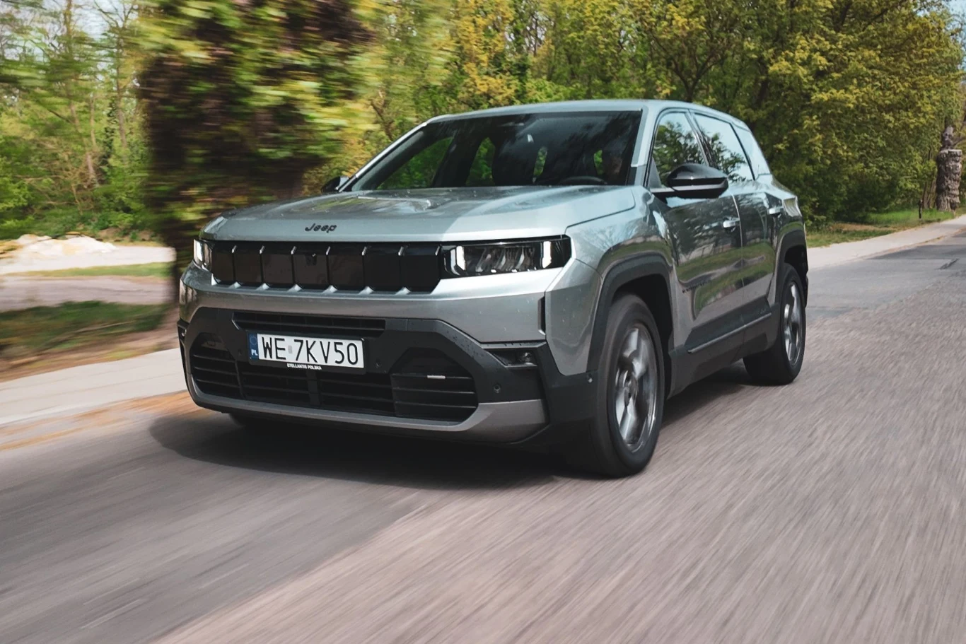 Jeep Compass z układem hybrydowym stara się jak najczęściej korzystać z napędu elektrycznego. Srebrny Jeep Compass jedzie po asfaltowej drodze otoczonej zielonymi drzewami.