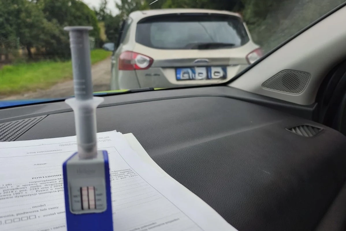 Policja stosuje zarówno jednorazowe narkotesty, jak i urządzenia wielokrotnego użytku Narkotester z pozytywnym wynikiem oraz dokumenty leżące na desce rozdzielczej w policyjnym radiowozie, zaparkowanym za srebrnym samochodem osobowym na bocznej, leśnej drodze.