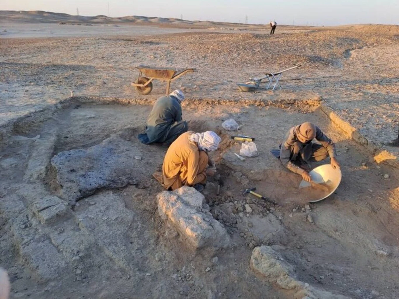 Archeolodzy podczas prac na stanowisku Shahr-i Sokhta. Trzech archeologów pracuje w wykopie na stanowisku pustynnym, wykorzystując narzędzia do odsłaniania i oczyszczania fragmentów kamiennych struktur.