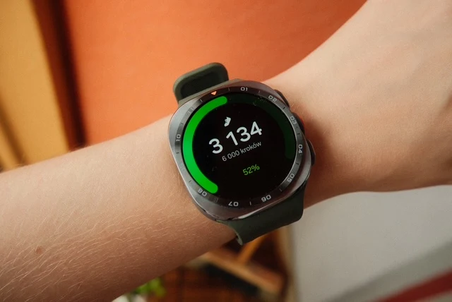 Czarny smartwatch na nadgarstku pokazujący liczbę 3 134 wykonanych kroków oraz postęp dziennego celu na kolorowym pasku wokół ekranu.
