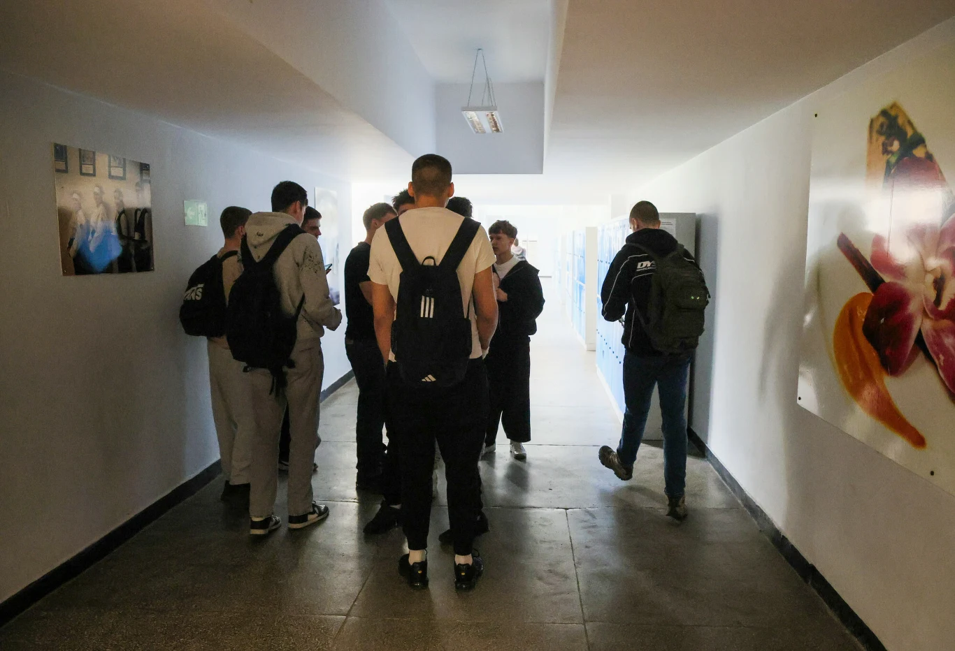 Grupa młodych osób z plecakami stojąca i rozmawiająca w jasnym szkolnym korytarzu.