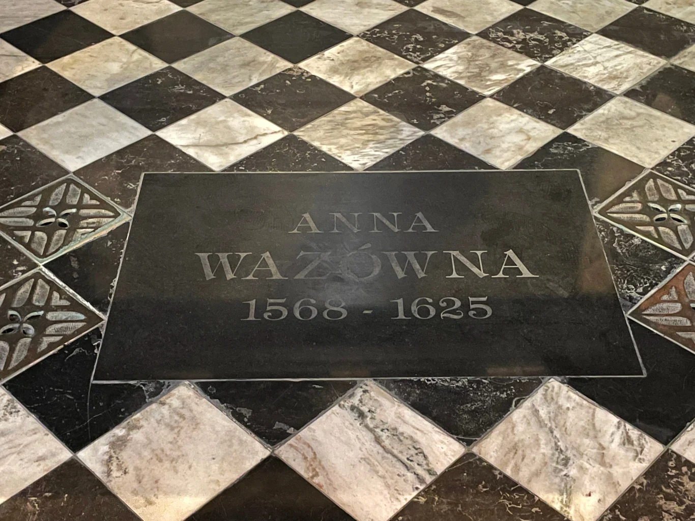 Kamienna płyta nagrobna z inskrypcją upamiętniającą Annę Wazównę, otoczona czarno-białą mozaiką posadzki z geometrycznym wzorem.