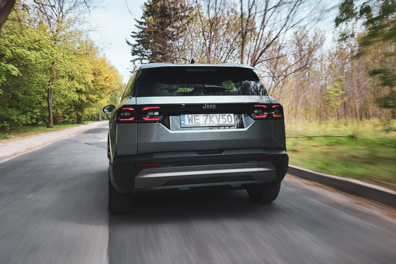 Silnik elektryczny zapewnia odpowiednie wsparcie jednostce. W efekcie dość sprawnie reaguje na gaz. Nowoczesny SUV marki Jeep porusza się asfaltową drogą otoczoną zielonymi i bezlistnymi drzewami.