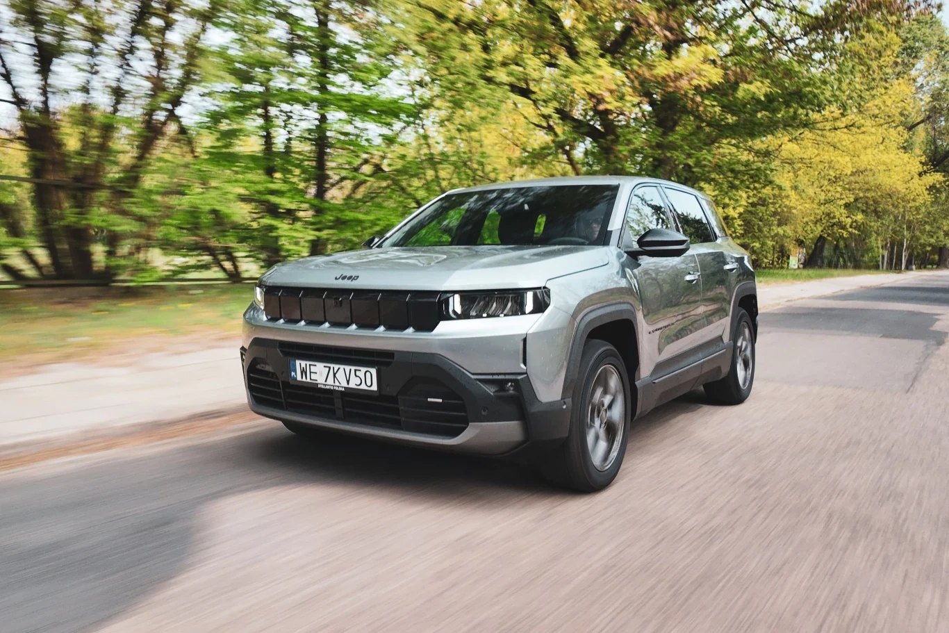 W podstawowej specyfikacji, podobnie jak w większości innych, Jeep Compass oferuje napęd na przód. AWD dostępne jest tylko w przypadku topowej odmiany elektrycznej. Srebrny SUV marki Jeep jadący po asfaltowej drodze otoczonej zielonymi drzewami.
