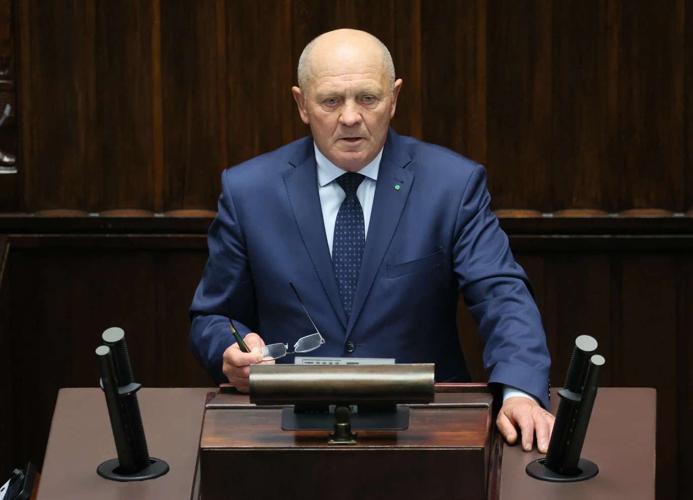 Mężczyzna w garniturze i krawacie stoi za mównicą, trzymając okulary w jednej ręce, na tle drewnianej ściany.