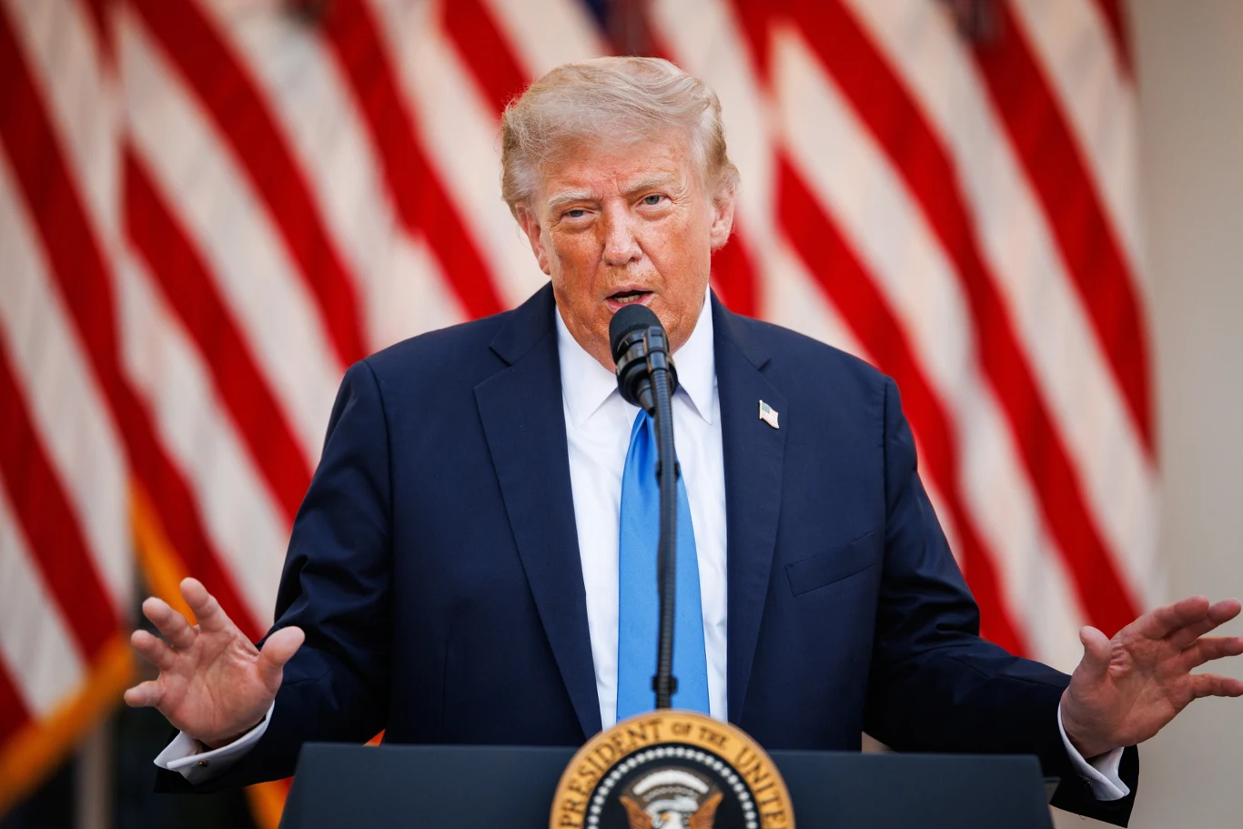 Prezydent Donlad Trump Donald Trump przemawia do mikrofonu na podium, w granatowym garniturze, na tle flag USA