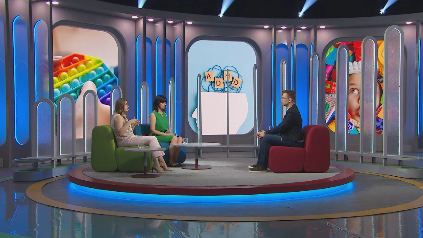 Trzy osoby siedzące na kolorowych fotelach podczas rozmowy w nowoczesnym studio telewizyjnym, za nimi widoczne duże ekrany z kolorowymi grafikami zabawek i dziecka.