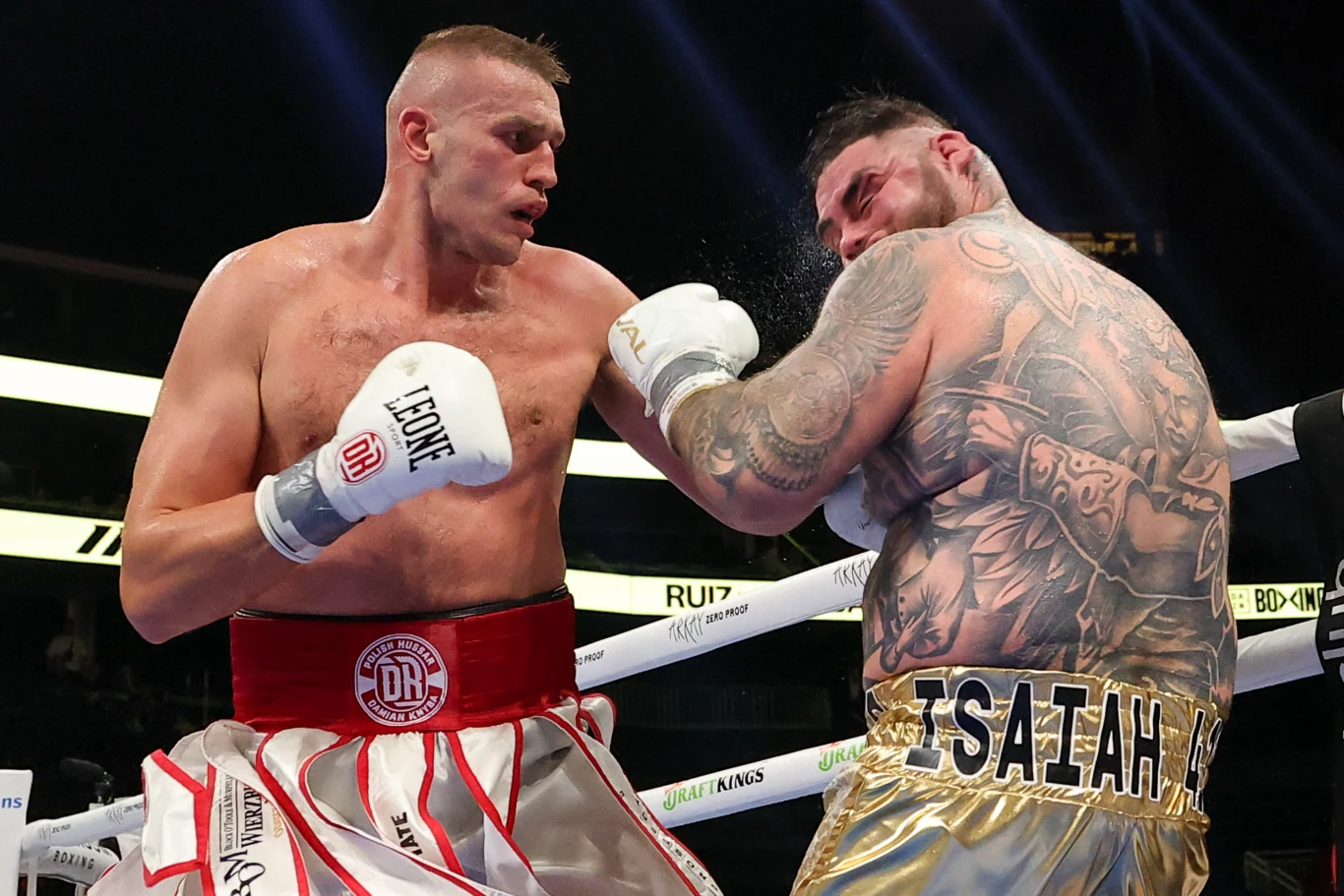 Damian Knyba i Andy Ruiz Jr. Dwaj bokserzy w trakcie intensywnej walki na ringu, jeden uderza drugiego mocnym ciosem w twarz, a wokół nich unosi się pot i pył z uderzenia.