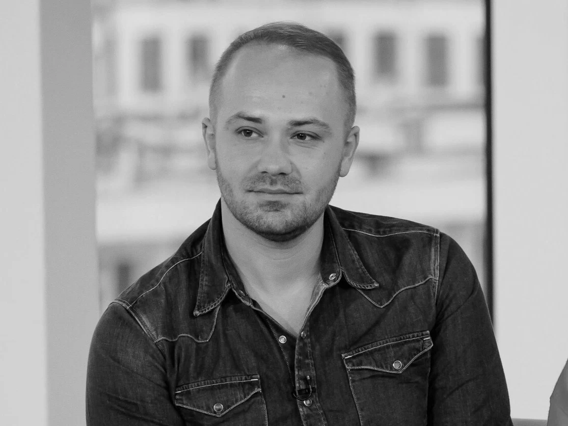 Adrian Szymaniak Adrian Szymaniak w ciemnej koszuli jeansowej, z krótkimi włosami i zarostem, siedzi przy oknie z rozmytym tłem.