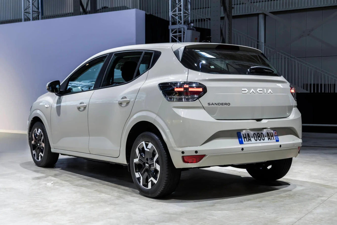 Jasnoszara Dacia Sandero nowej generacji z przyciemnianymi szybami, czarnymi detalami nadwozia oraz charakterystycznymi tylnymi światłami LED, ustawiona na jasnej posadzce w jasnym, industrialnym otoczeniu.