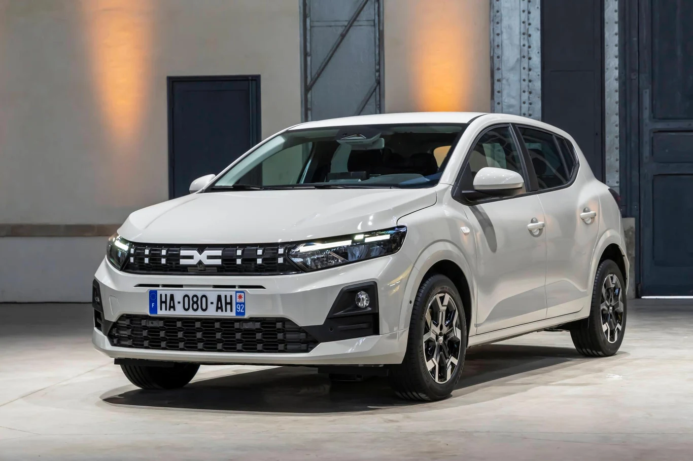 Biały samochód osobowy Dacia Sandero drugiej generacji stoi na betonowej podłodze, wyeksponowany w jasnym, industrialnym wnętrzu z metalowymi drzwiami w tle.