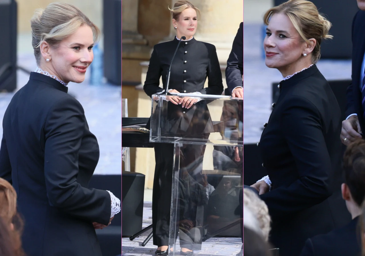 Kobieta w eleganckim czarnym garniturze ze srebrnymi guzikami i koronkowym wykończeniem mankietów przemawia przy mównicy przed zgromadzonymi ludźmi, zwracając uwagę profesjonalnym, stonowanym wyglądem.