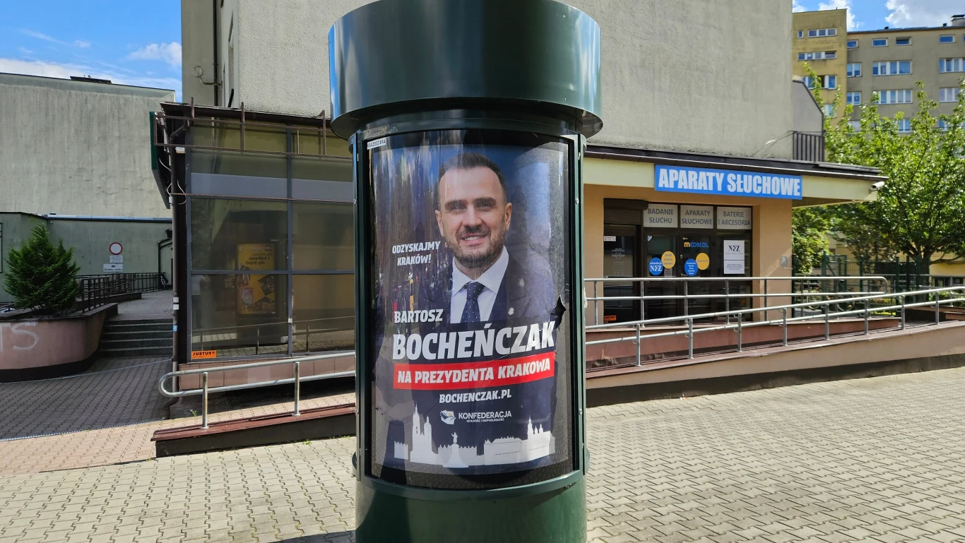 Plakat wyborczy Bartosza Bochenčzaka z wizerunkiem kandydata oraz napisem informującym o jego kandydaturze na prezydenta Krajowej, umieszczony na słupie ogłoszeniowym na tle budynku z szyldem 'Aparaty słuchowe'.