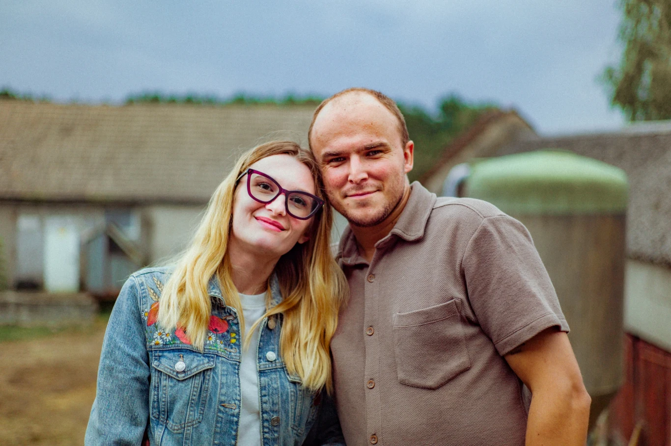 Kobieta w okularach i dżinsowej kurtce stoi blisko uśmiechniętego mężczyzny w beżowej koszuli, oboje patrzą wprost do obiektywu.
