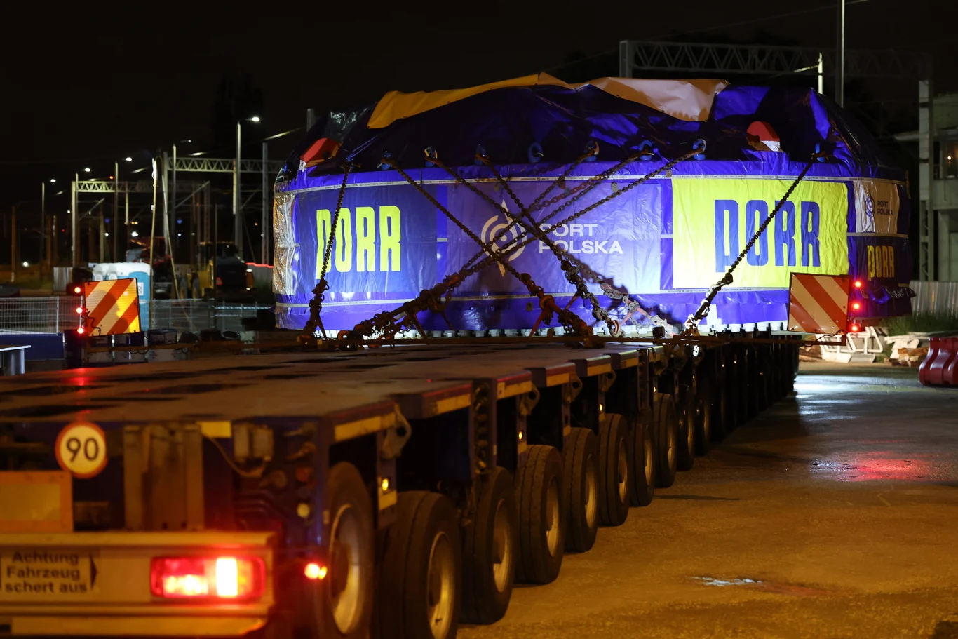 Sam napęd maszyny TBM waży 169 ton. Cały transport - 270 ton Ogromny ładunek owinięty niebieską plandeką z żółtym napisem „DORR” transportowany na wieloosiowej platformie, otoczony zabezpieczeniami i oświetleniem nocnym.
