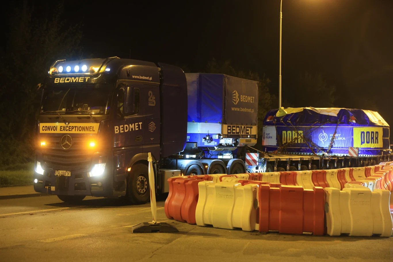 Niskopodłowowa laweta z napędem TBM była podpięta do czteroosiowego ciągnika balastowego Ciemnoniebieska ciężarówka z naczepą typu platforma przewozi duży ładunek owinięty niebieską plandeką, osłonięta barierkami zabezpieczającymi, na oświetlonym odcinku drogi w nocy.