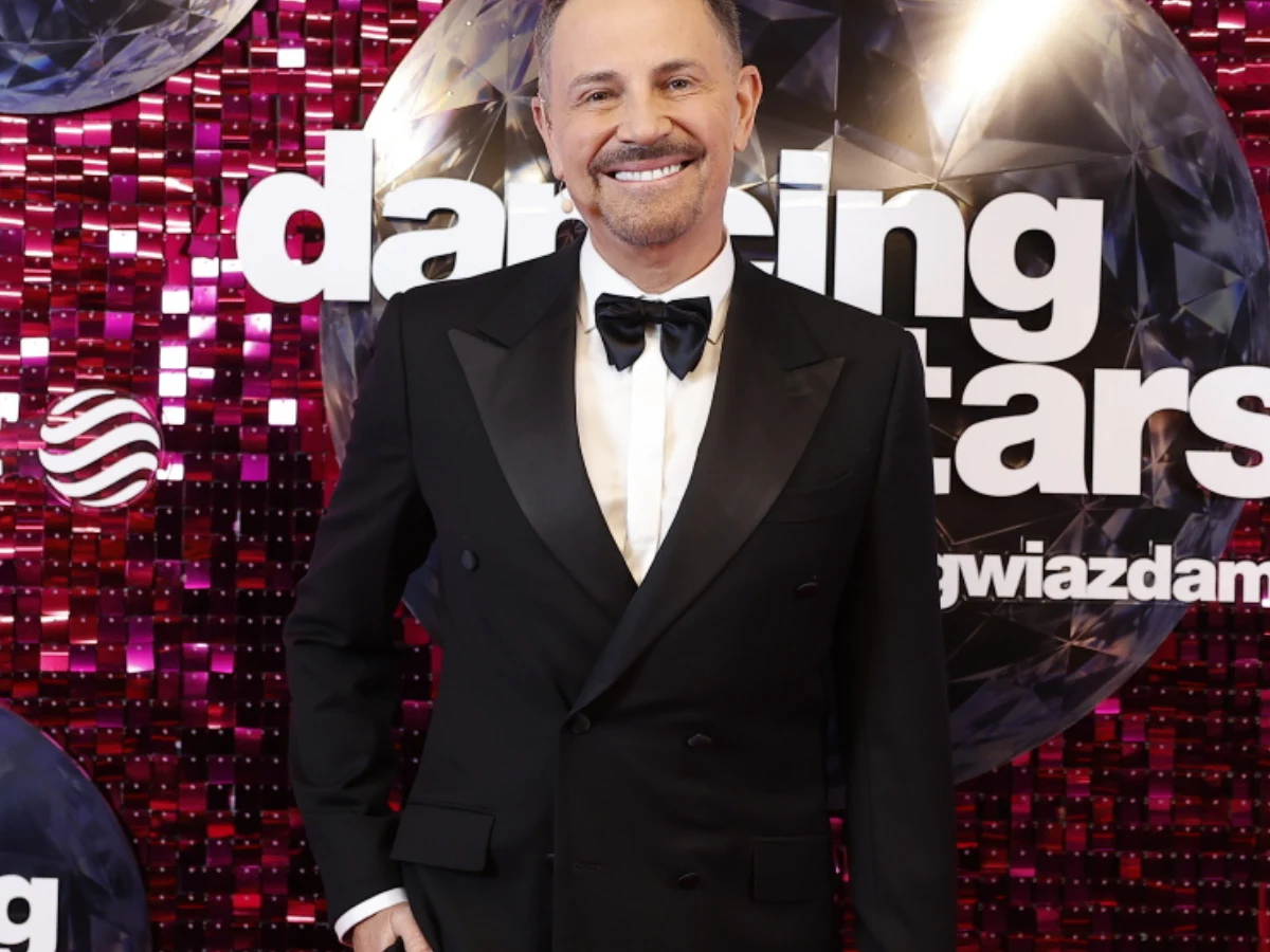 Elegancko ubrany mężczyzna w czarnym smokingu i muszce stoi na tle ścianki z logo programu 'Dancing with the Stars'.