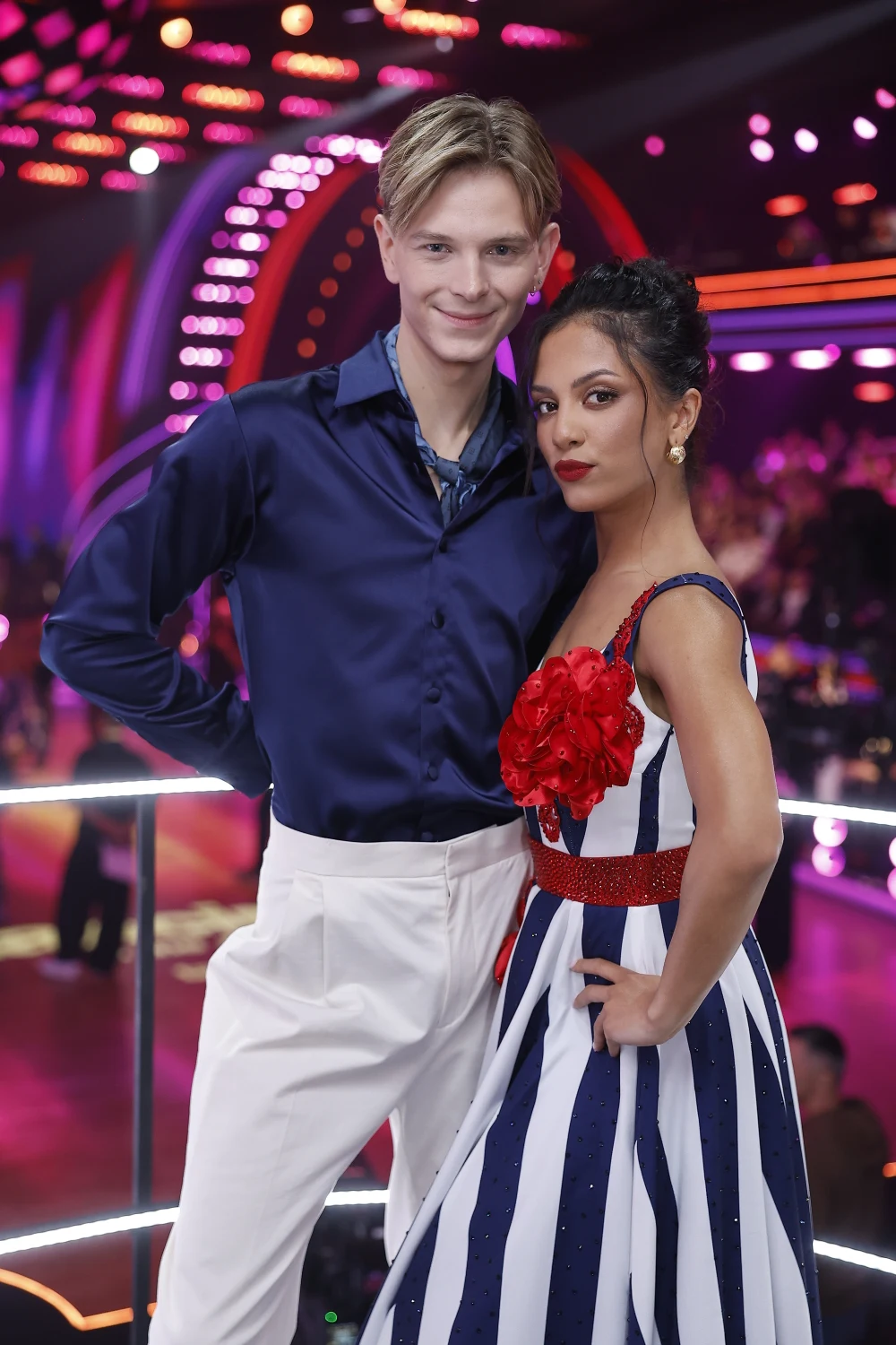 Dominik Rupiński i Estelle Français Para ubrana w eleganckie ubrania taneczne, mężczyzna w ciemnoniebieskiej koszuli i białych spodniach, kobieta w biało-granatowej sukni w paski z czerwonymi kwiatami na piersi, w tle kolorowe światła sceniczne i rozmyta publiczność.