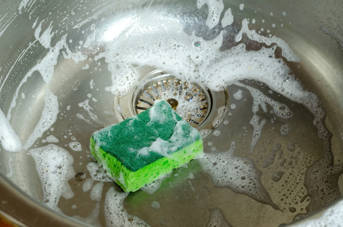 Aby problemy z nieprzyjemnym zapachem w kuchni zdarzały się rzadziej, warto regularnie czyścić syfon kuchenny. zielona gąbka do mycia naczyń pokryta pianą leży w metalowym zlewie z resztkami wody i piany wokół odpływu