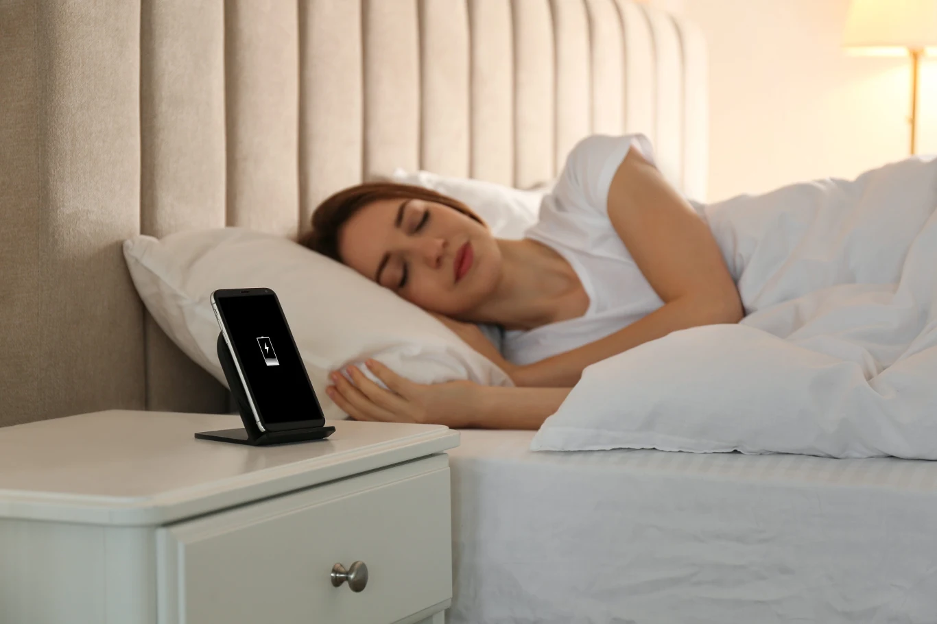 Co robi smartfon w trakcie nocnego ładowania? To często dzieje się w tle. Kobieta śpi w łóżku obok ładowanego na nocnej szafce smartfona na podstawce bezprzewodowej.