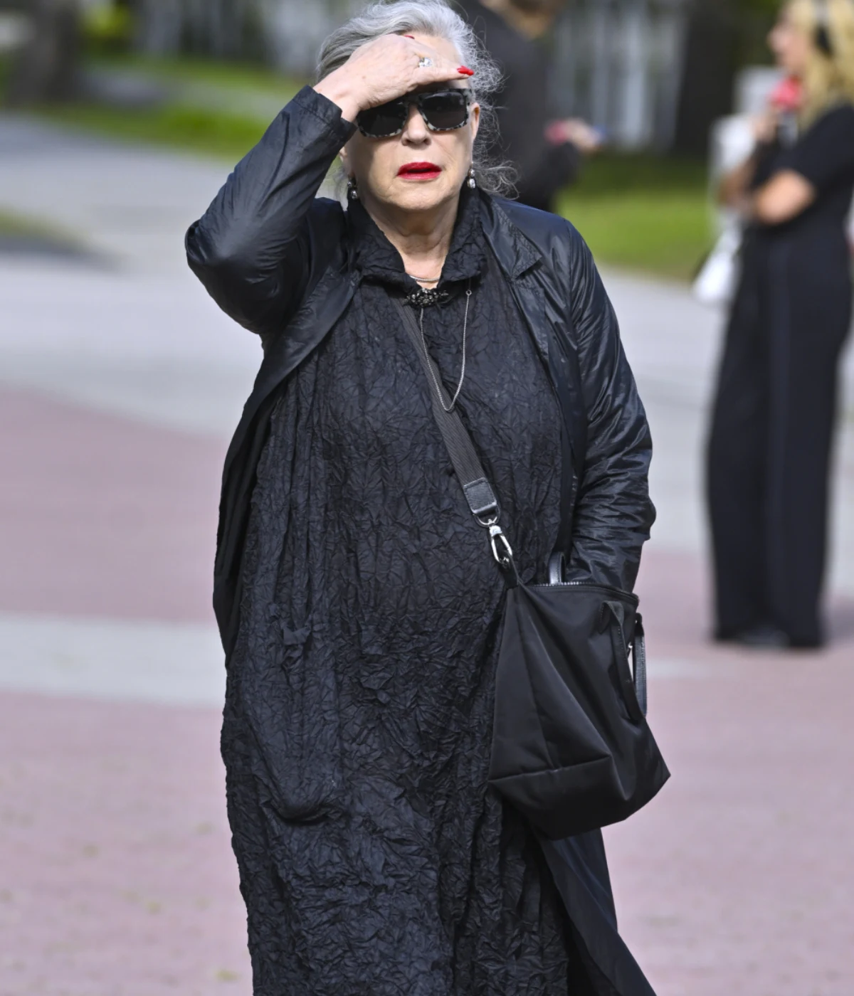 Krystyna Janda Starsza kobieta z siwymi włosami spiętymi do tyłu, ubrana w czarną, luźną sukienkę i ciemne okulary przeciwsłoneczne, trzyma rękę na czole, z czarną torbą przewieszoną przez ramię.
