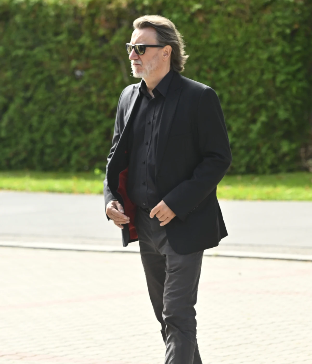 Robert Więckiewicz Mężczyzna w ciemnym garniturze i okularach przeciwsłonecznych idzie chodnikiem w otoczeniu zieleni.