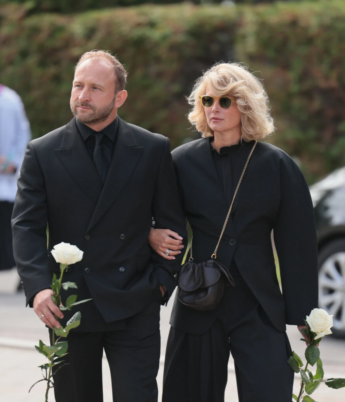 Borys Szyc i Justyna Szyc-Nagłowska Elegancko ubrany mężczyzna i kobieta w czarnych strojach idą razem, trzymając w dłoniach białe róże.