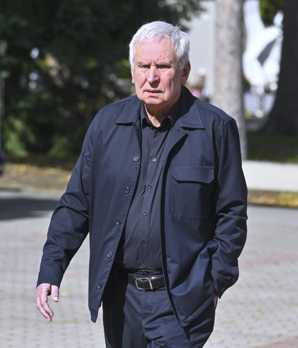 Krzysztof Materna Starszy mężczyzna o siwych włosach ubrany w ciemny garnitur i koszulę idzie chodnikiem w słoneczny dzień, trzymając jedną rękę w kieszeni.