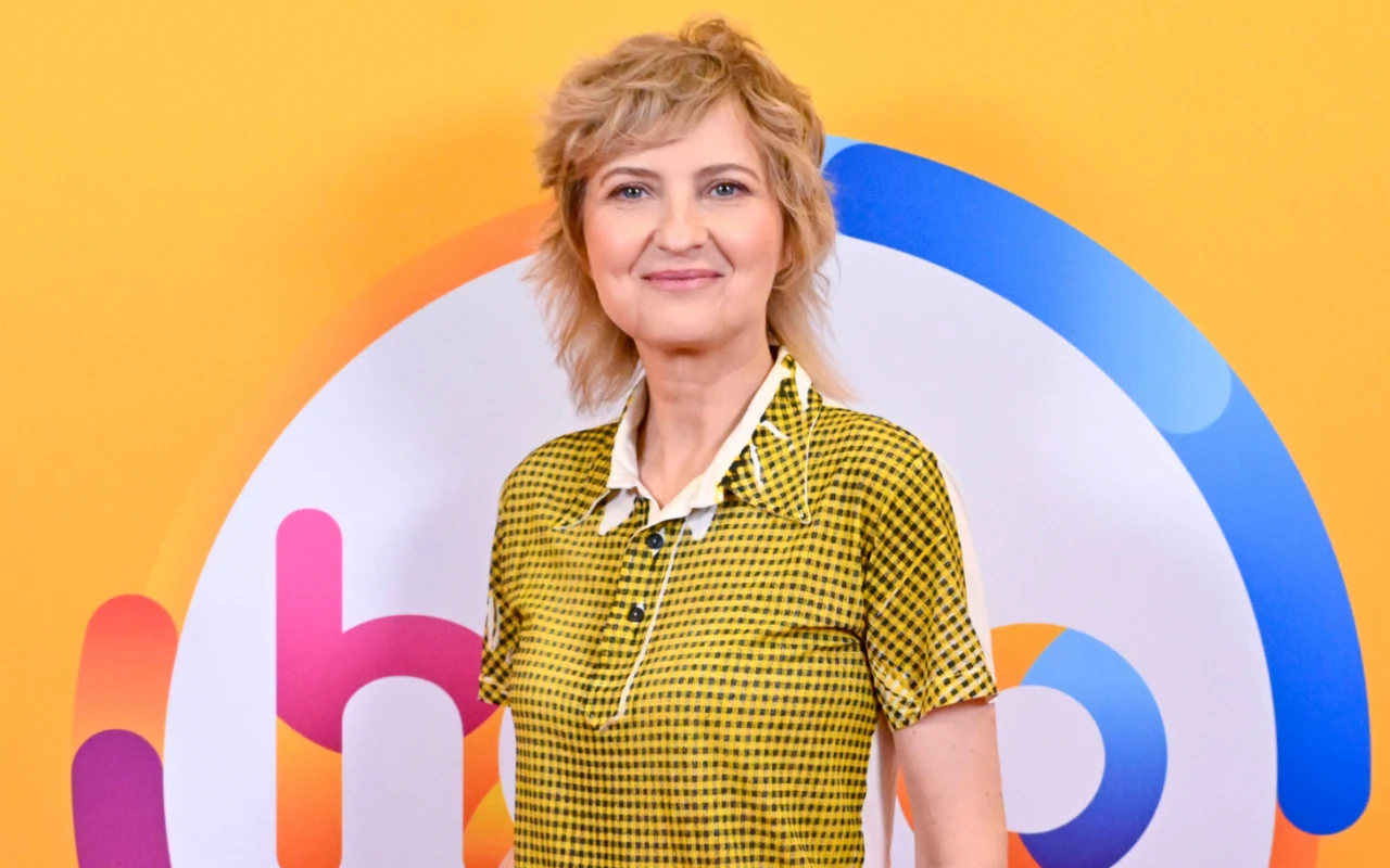 Izabela Kuna w studiu "halo tu polsat" Kobieta z krótkimi blond włosami, ubrana w wzorzystą koszulę w odcieniach żółci, stoi na tle kolorowego graficznego logo.