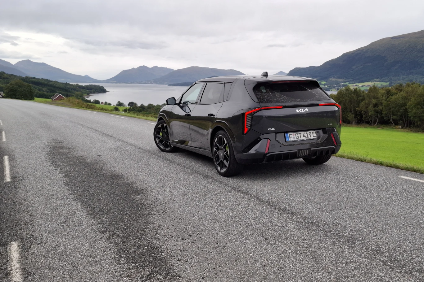Kia EV4 GT Czarny samochód marki Kia stoi na pustej asfaltowej drodze z widokiem na jezioro i otaczające je zielone wzgórza oraz góry.