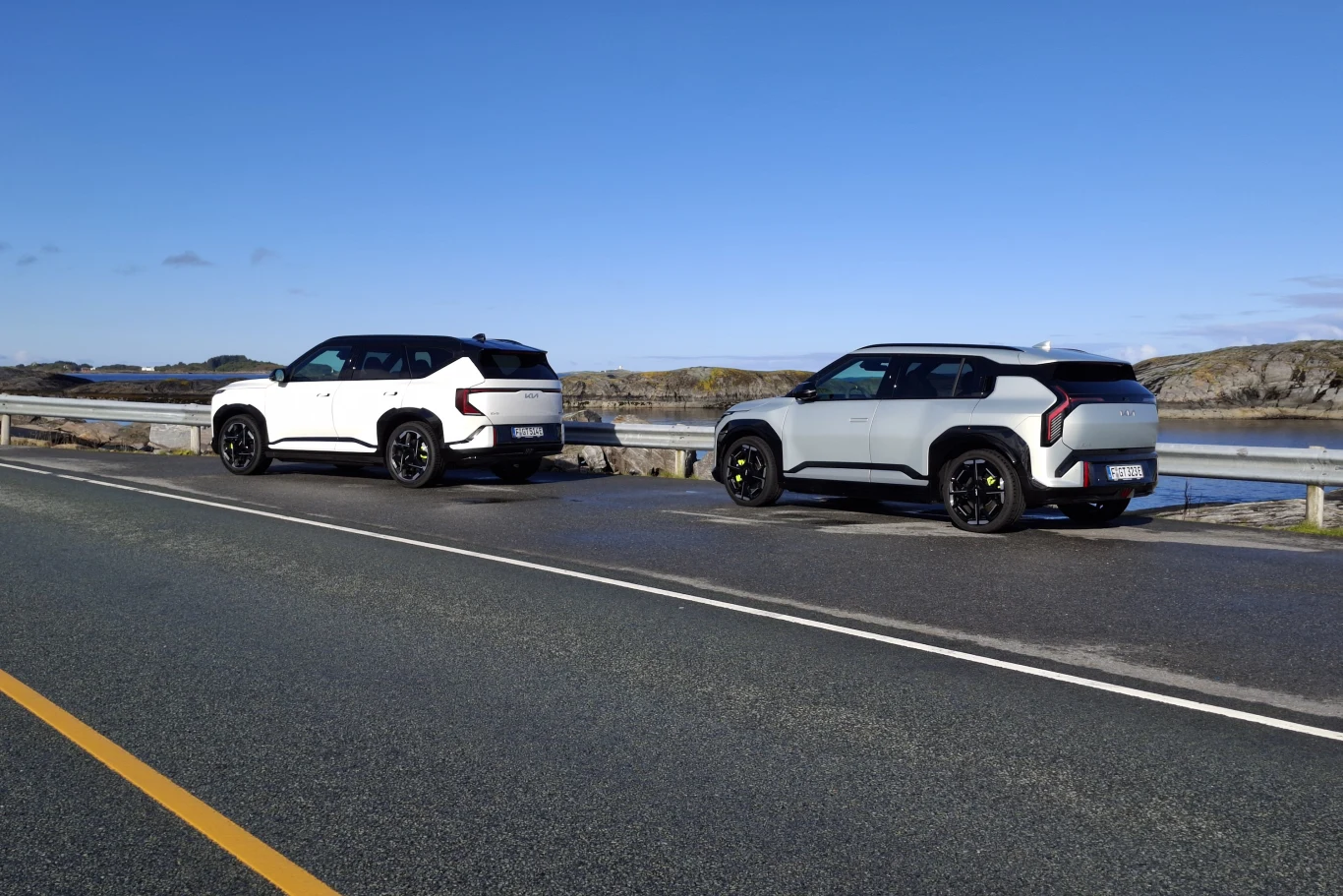 Kia EV5 GT i Kia EV3 GT Dwa nowoczesne SUV-y o jasnej karoserii zaparkowane na poboczu asfaltowej drogi przy nadmorskiej scenerii, z widocznym otwartym pejzażem w tle.