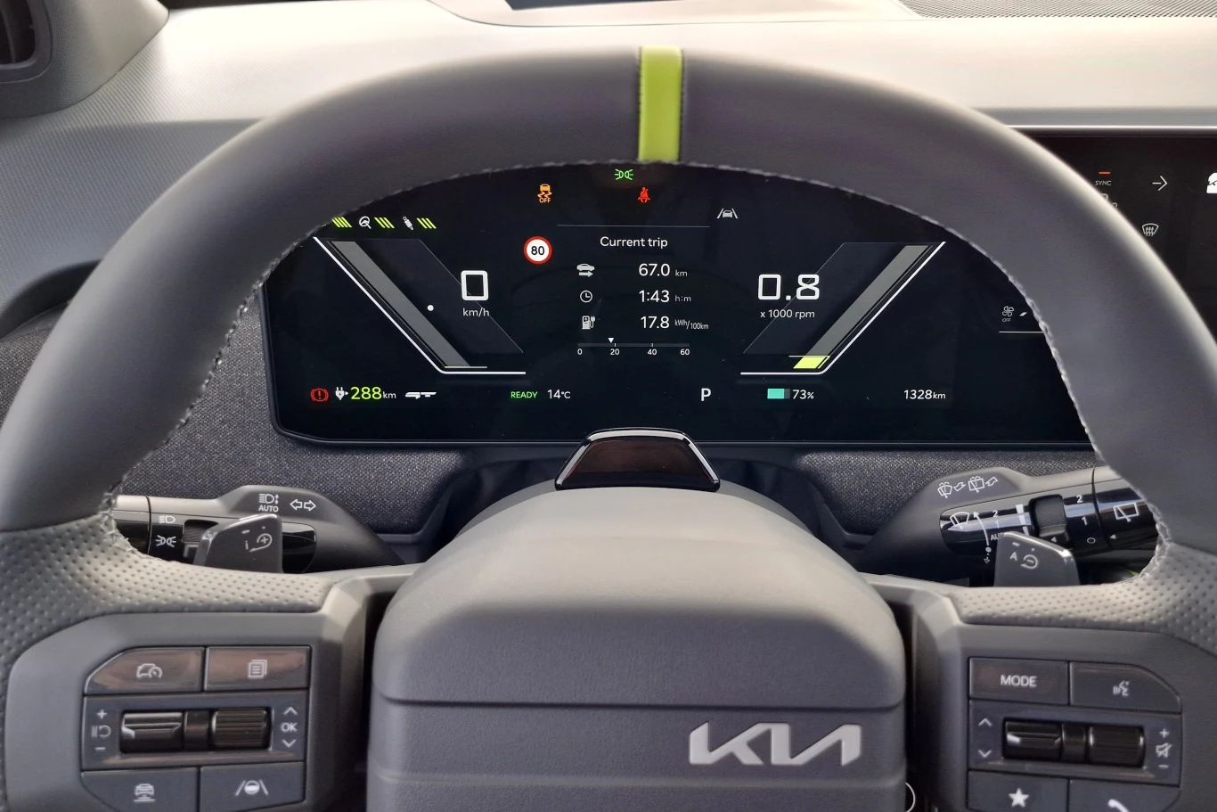 Kia EV3 GT Cyfrowy kokpit samochodu marki Kia z wyświetlaczem prezentującym aktualną prędkość, stan baterii, zasięg oraz informacje dotyczące jazdy, otoczony nowoczesną kierownicą z wbudowanymi przyciskami sterującymi.