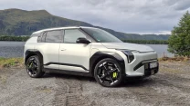 Nowoczesny SUV o geometrycznych kształtach parkuje na żwirowym podłożu, z malowniczym jeziorem i górzystym terenem w tle.