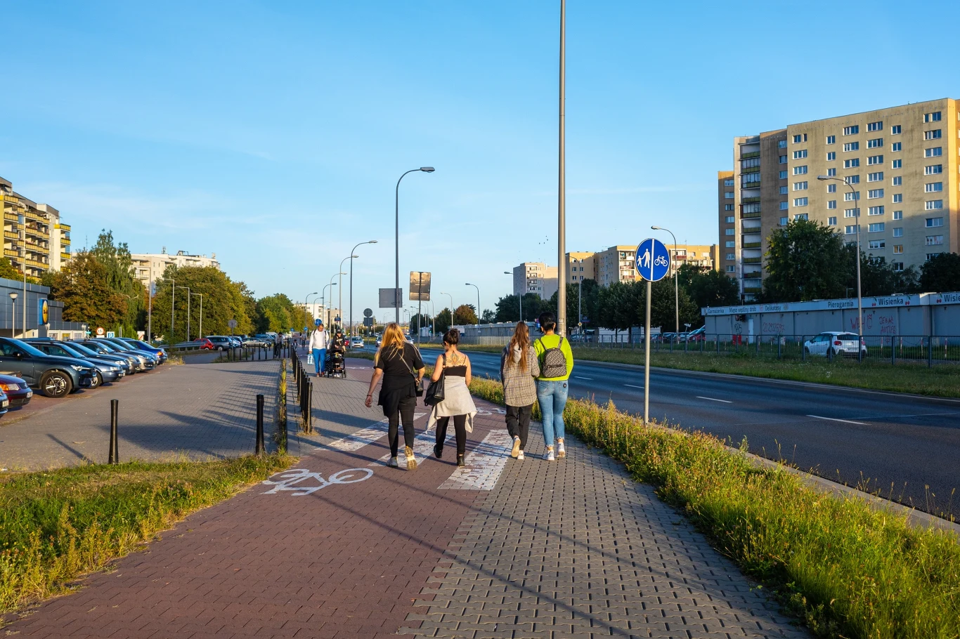 Zebra na ścieżce rowerowej. Czy pieszy ma pierwszeństwo? Zebra na drodze rowerowej, osoby spacerujące chodnikiem przy ulicy z blokami i zaparkowanymi samochodami.