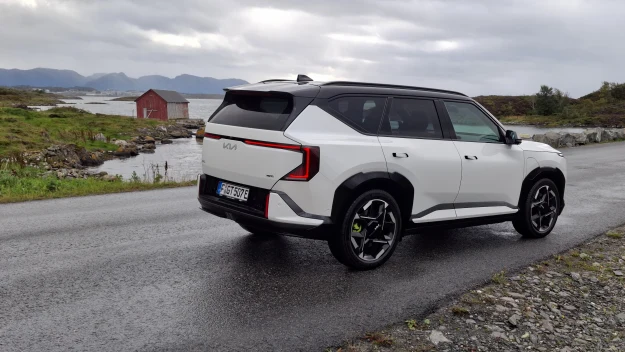 Biały, nowoczesny SUV marki Kia z czarnymi elementami nadwozia stoi na mokrej drodze asfaltowej, w tle surowy krajobraz z jeziorem i drewnianym, czerwonym domkiem.