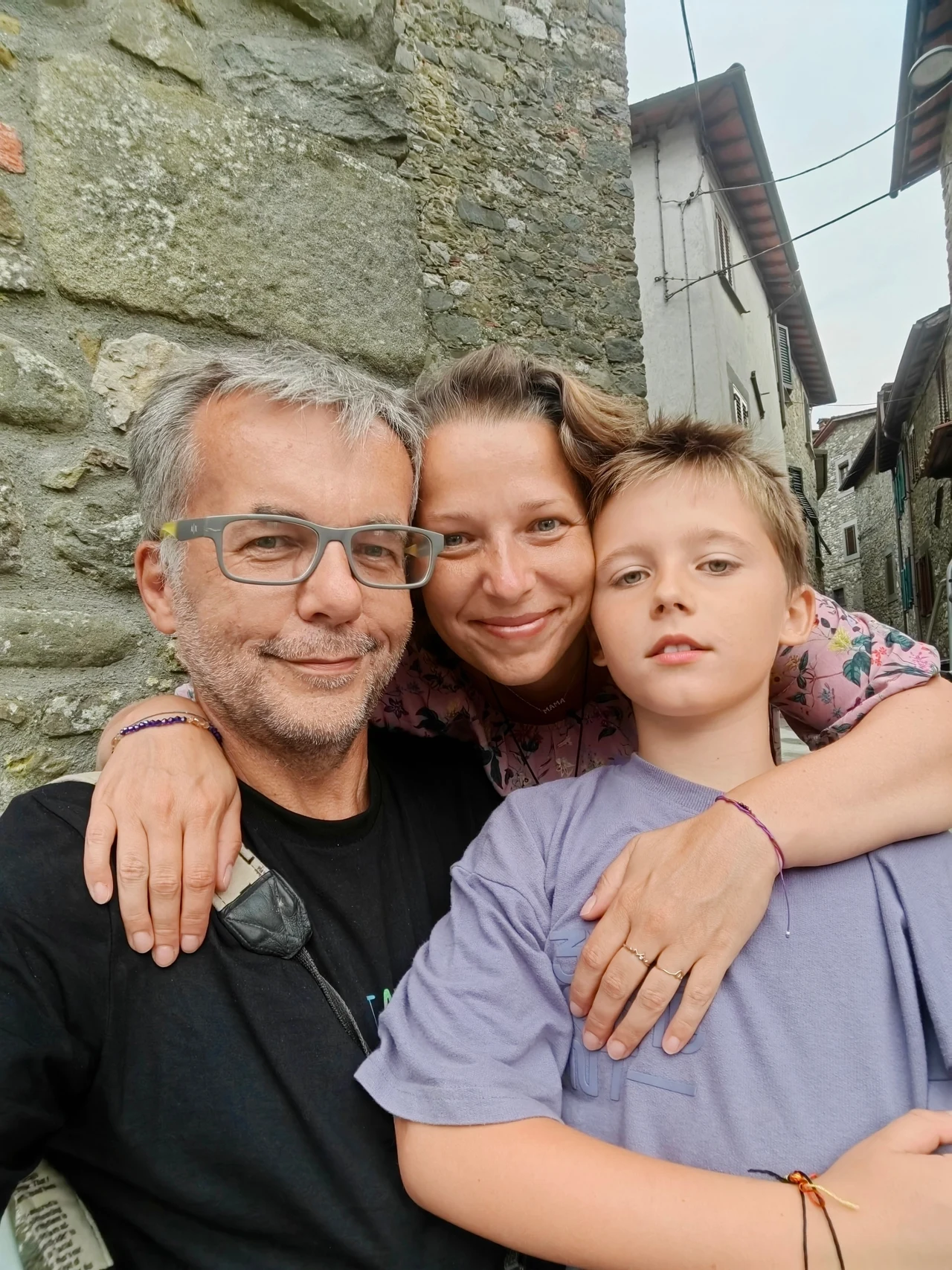 Trzyosobowa rodzina obejmująca się i uśmiechająca do aparatu, stojąca przy kamiennej ścianie w wąskiej, historycznej uliczce.