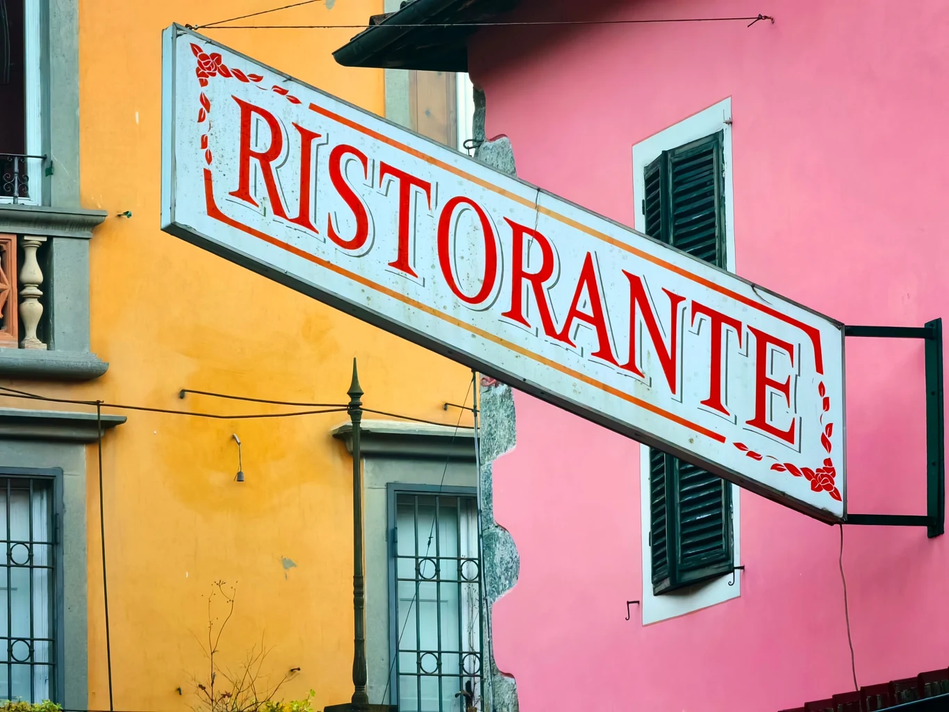 Duży biały szyld z czerwonym napisem „RISTORANTE” zamocowany na ścianie różowego budynku z zielonym oknem i żaluzją, w tle żółty budynek z oknami.