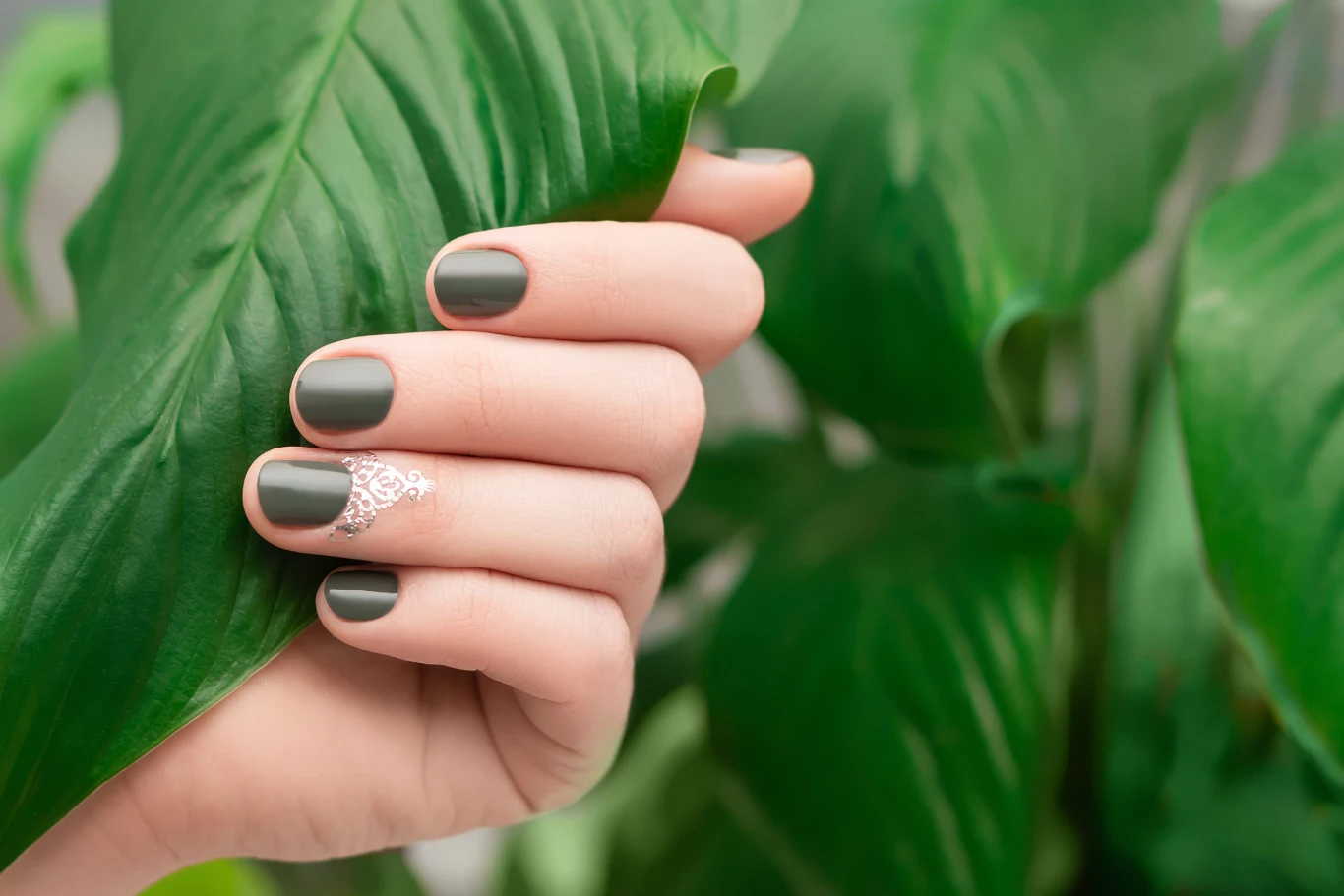 Dłoń z zielonym manicure oraz ozdobnym białym wzorem na jednym paznokciu przytrzymująca duży zielony liść.