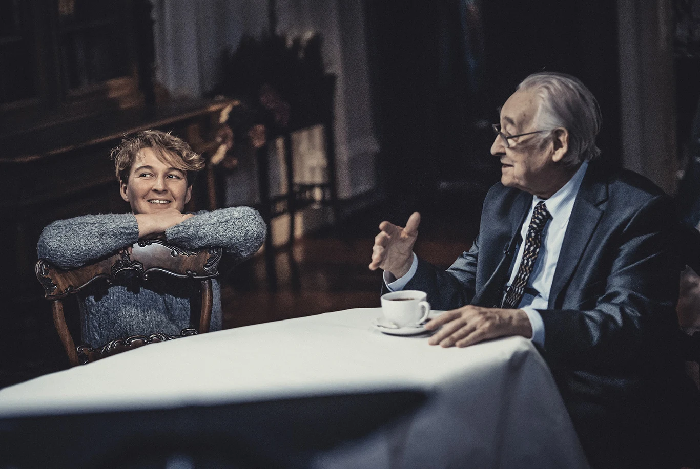 Joanna Rożen-Wojciechowska i Andrzej Wajda / Centrum Kultury Filmowej im. Andrzeja Wajdy Starszy mężczyzna w garniturze siedzi przy stole z filiżanką kawy, rozmawiając z uśmiechniętą kobietą opartą swobodnie o krzesło obok.