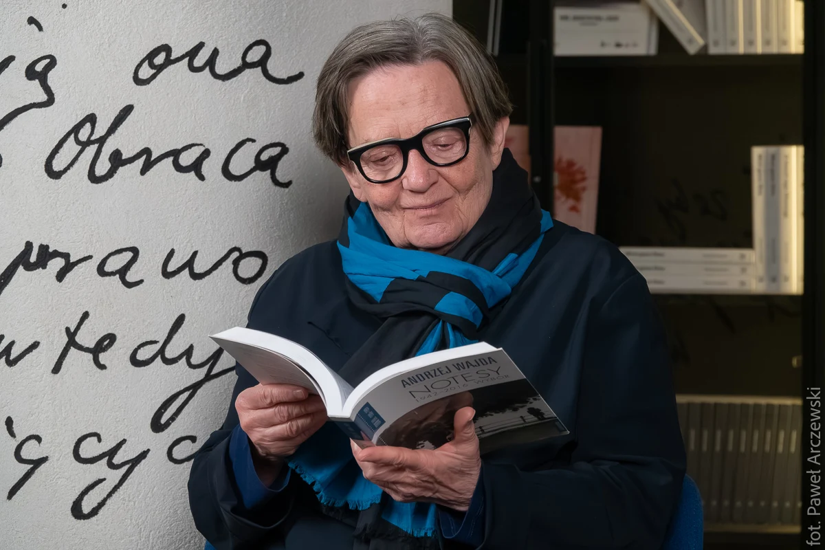 Agnieszka Holland czyta "Notesy" Andrzeja Wajdy / Centrum Kultury Filmowej im. Andrzeja Wajdy Starsza osoba w okularach i niebieskim szaliku siedzi z otwartą książką, uśmiechając się podczas lektury, w tle ściana z dużymi, czarnymi napisami oraz półka z książkami.