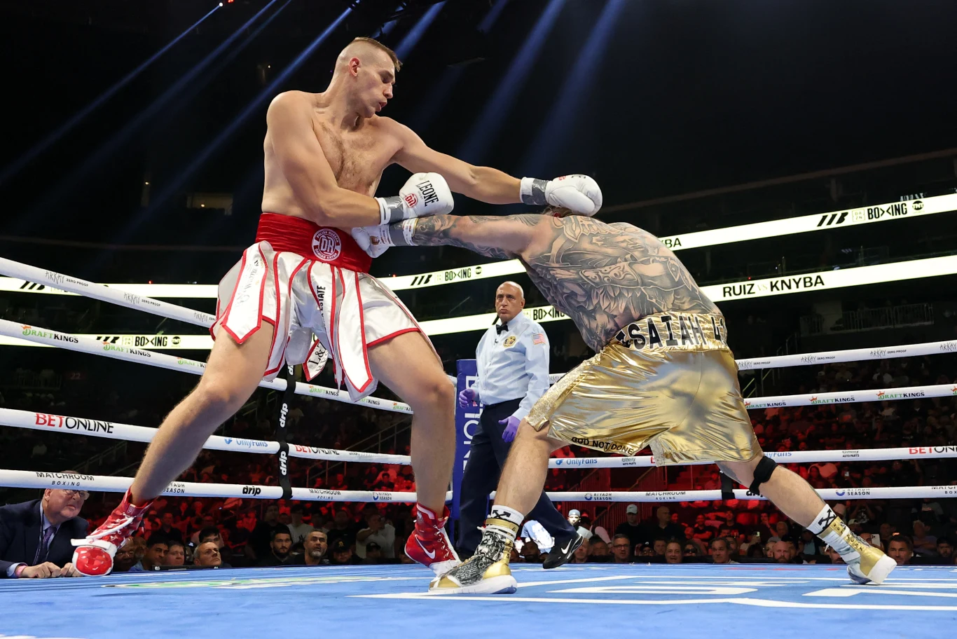 Damian Knyba - Andy Ruiz jr Dwóch bokserów wymienia ciosy w ringu, jeden ubrany w czerwone spodenki zadaje prosty lewą ręką, drugi w złotych szortach pochyla się, unikając uderzenia; w tle sędzia obserwuje walkę.