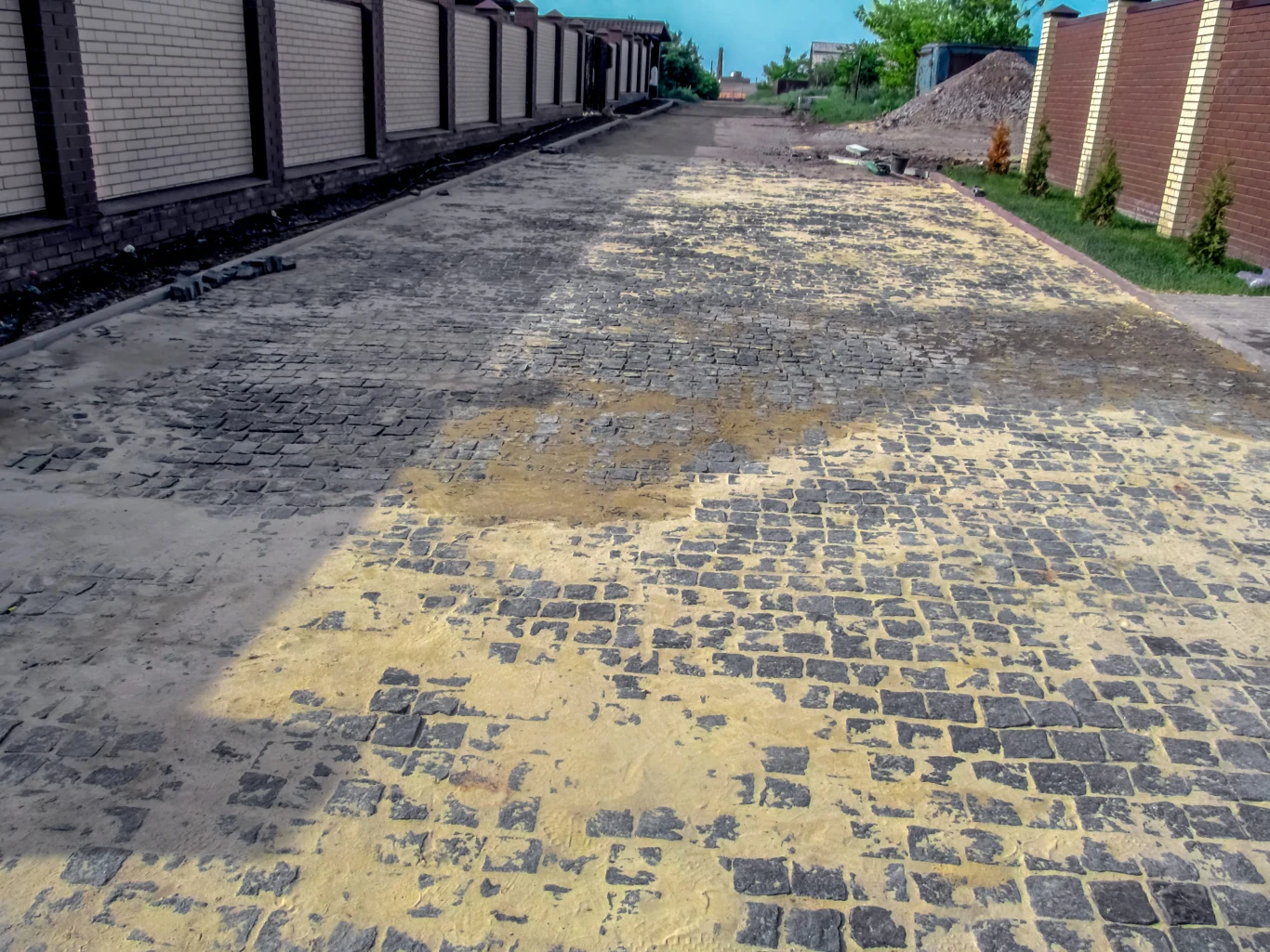 Po myciu i czyszczeniu trzeba uzupełnić przestrzenie między kostkami Brukowana droga pomiędzy wysokimi, murowanymi ogrodzeniami, z widocznym rozsypanym piaskiem na powierzchni kostki brukowej.